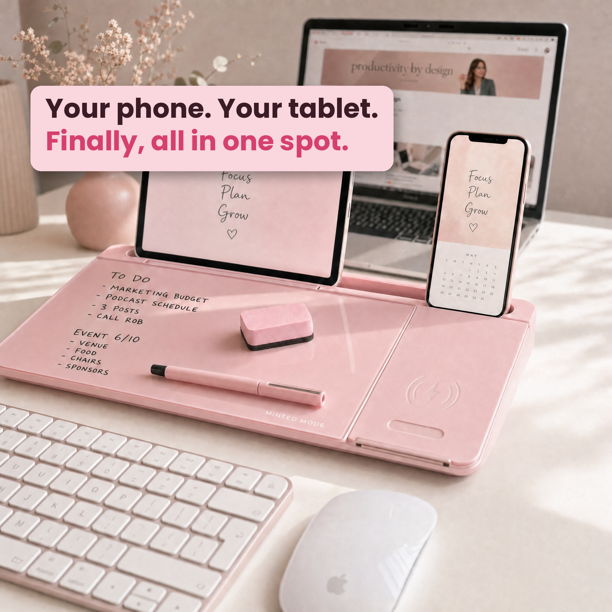 A pink all-in-one desk organizer and charging station shown on a light tabletop, with a tablet propped in the center, a phone docked on the right, a keyboard and mouse in front, and a laptop in the background. A pink text bubble reads, 'Your phone.